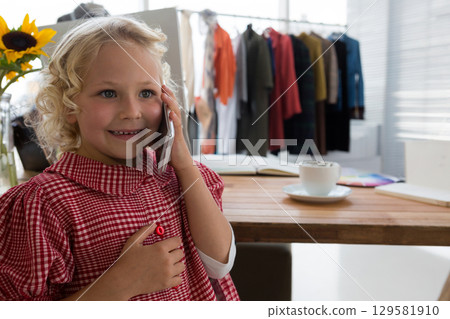 Girl talking on smartphone in home office at table with sunflower and clothing rack, copy space 129581910