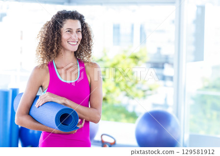 Woman holding rolled blue yoga mat at gym, wearing fuchsia tank top and leggings with kettlebell Woman holding rolled blue yoga mat at gym, wearing fuchsia tank top and leggings with kettlebell 129581912
