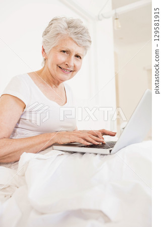 Senior woman sitting up in bed smiling while using laptop under hanging light fixture in bedroom 129581915