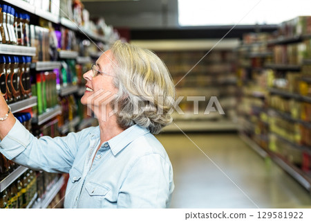 Senior female shopper reaching up selecting sauce bottle on supermarket aisle shelves, copy space 129581922