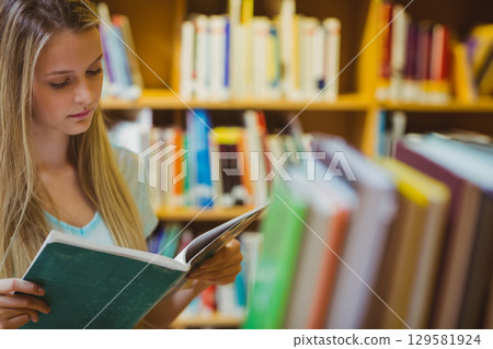 Teenage girl reading open book in library aisle with bookshelves and blurred books, copy space Teenage girl reading open book in library aisle with bookshelves and blurred books, copy space 129581924