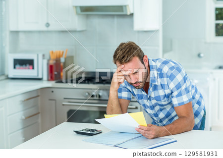 Middle-aged man leaning at kitchen island, holding papers, reviewing folder, using calculator 129581931