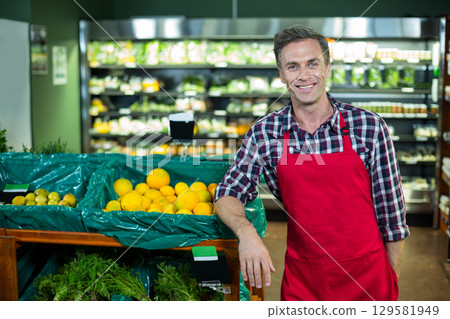 Middle-aged man in apron leaning on bin at supermarket produce aisle showing lemons with price sign Middle-aged man in apron leaning on bin at supermarket produce aisle showing lemons with price sign 129581949