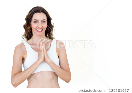 Woman smiling and holding palms together in prayer pose in studio wearing white sports bra 129581957