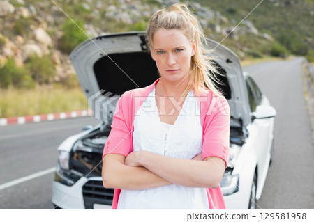 Woman in pink cardigan standing with arms crossed by white sedan hood open on mountain road 129581958