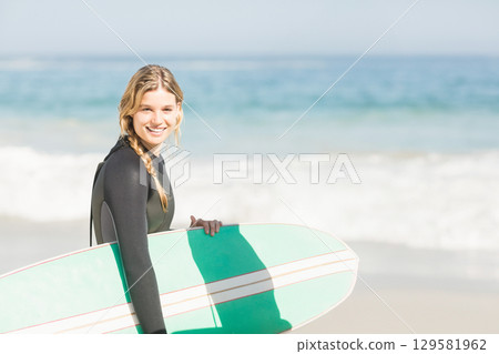 Female surfer holding green-white surfboard standing in wetsuit on sand by gentle waves, copy space 129581962