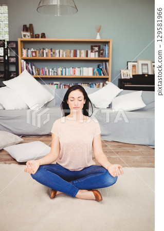 Woman sitting cross-legged meditating on beige rug in living room with bookshelf 129581966