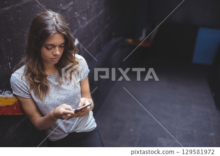 Woman mid-20s leaning against painted wall in urban alley holding smartphone near ledge, copy space 129581972