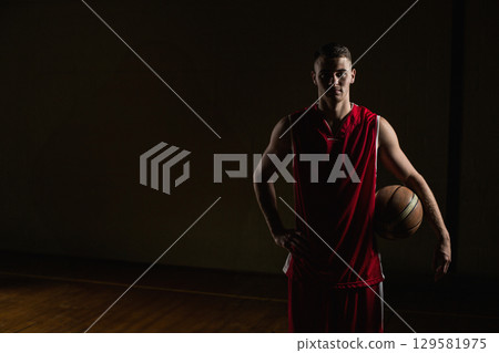 Male basketball player wearing red uniform holding basketball at side standing on court in gym 129581975