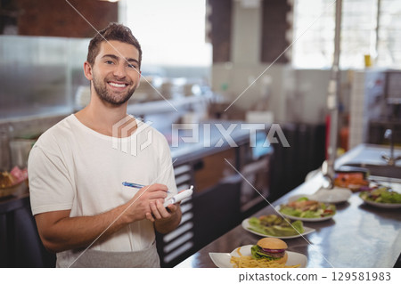 Male server in apron smiling and holding notepad and pen at steel kitchen counter, copy space 129581983