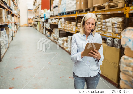 Woman standing in warehouse aisle scanning inventory on clipboard and wearing headset, copy space Woman standing in warehouse aisle scanning inventory on clipboard and wearing headset, copy space 129581994