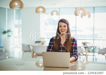 Woman wearing plaid shirt working on laptop at modern office with wooden desk, pendant lamps, plant Woman wearing plaid shirt working on laptop at modern office with wooden desk, pendant lamps, plant 129581997