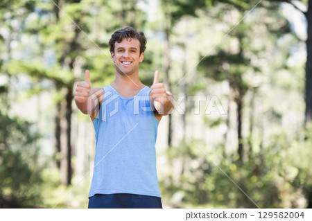 Smiling man extending arms with thumbs up in sunlit forest clearing wearing athletic wear Smiling man extending arms with thumbs up in sunlit forest clearing wearing athletic wear 129582004