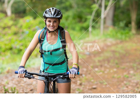 Female cyclist riding mountain bike on forest trail wearing black helmet carrying hydration pack 129582005