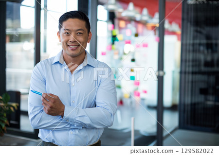 Asian man standing in modern open office holding blue pen near glass wall with sticky notes 129582022