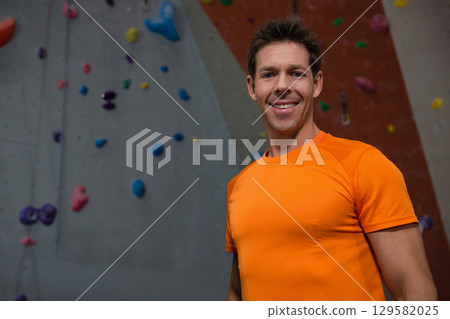 Mid adult man posing by climbing wall wearing orange shirt with holds and rope, copy space 129582025
