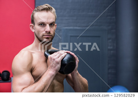 Shirtless muscular man lifting black kettlebell at chest height in gym area with dumbbell rack 129582029