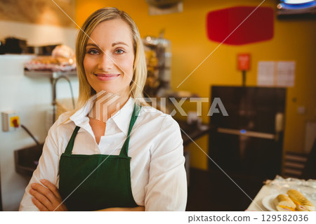 Female bakery staff wearing green apron displaying croissants and bread baskets at bakery counter 129582036