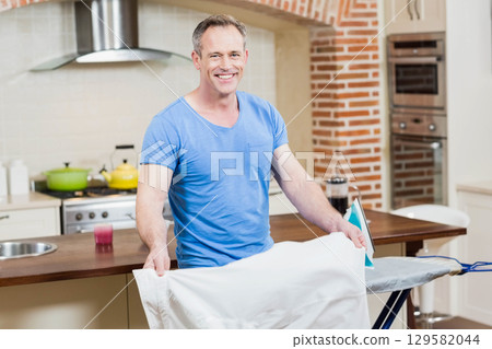 Senior man ironing white shirt with steam iron on ironing board in home kitchen with pots 129582044
