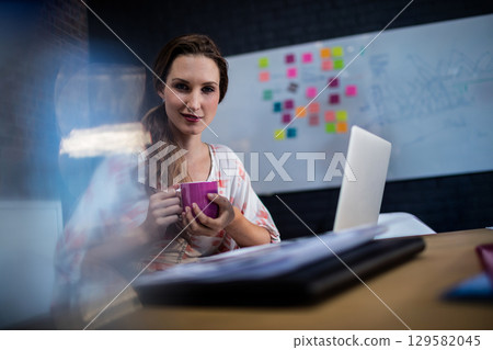 Female worker sitting at desk in meeting room, holding pink mug near silver laptop under whiteboard Female worker sitting at desk in meeting room, holding pink mug near silver laptop under whiteboard 129582045