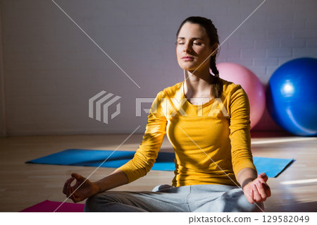 Woman in yellow top, gray sweatpants meditating on blue mat in studio next to exercise balls Woman in yellow top, gray sweatpants meditating on blue mat in studio next to exercise balls 129582049