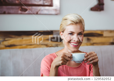 Woman wearing coral blouse smiling and holding white ceramic cup on café bench by wood-paneled wall Woman wearing coral blouse smiling and holding white ceramic cup on café bench by wood-paneled wall 129582052