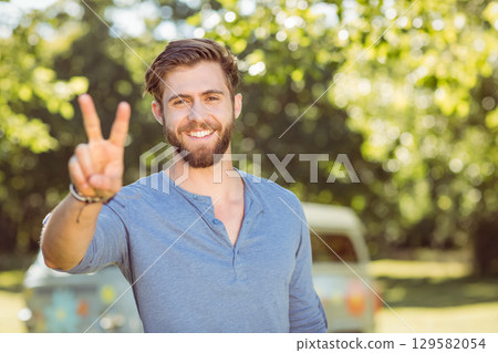 Man standing at grassy park wearing henley shirt with bracelets showing peace sign with camper van 129582054