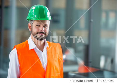 Man standing in site office wearing green hard hat and orange safety vest against glass partitions 129582060
