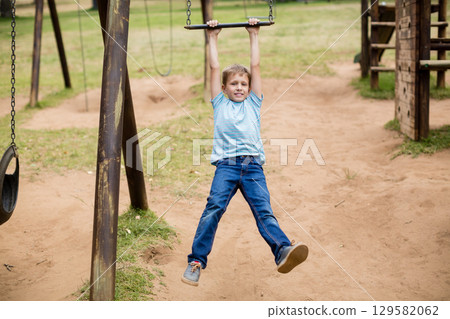 School-age boy hanging from horizontal bar on playground with tire swing and wooden climbing frame 129582062