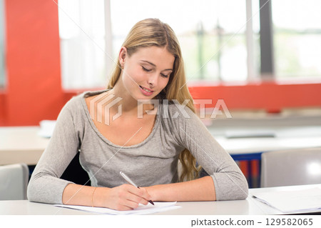 Teenage female student writing on paper with ballpoint pen at desk in classroom with red wall 129582065