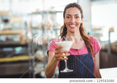 Mid adult female bartender in dark striped apron extending frothy martini glass at kitchen shelves Mid adult female bartender in dark striped apron extending frothy martini glass at kitchen shelves 129582080