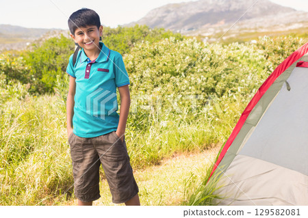 Boy standing next to camping tent with red trim in grassy meadow with shrubs, copy space 129582081
