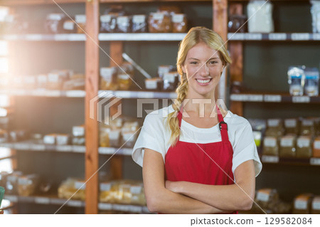 Female clerk standing with arms crossed in shop by wooden shelves with bulk jars and scanner Female clerk standing with arms crossed in shop by wooden shelves with bulk jars and scanner 129582084