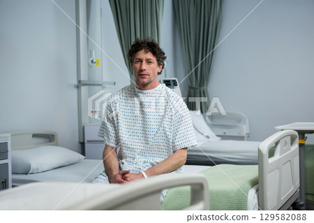 Male patient wearing hospital gown sitting on bed in ward beside IV stand and privacy curtains Male patient wearing hospital gown sitting on bed in ward beside IV stand and privacy curtains 129582088