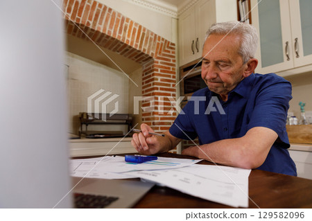 Senior man wearing navy-blue shirt reviewing documents on kitchen table with calculator, copy space 129582096