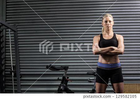 Female athlete standing with arms crossed at gym with metal shutter, bike, weight rack, copy space Female athlete standing with arms crossed at gym with metal shutter, bike, weight rack, copy space 129582105