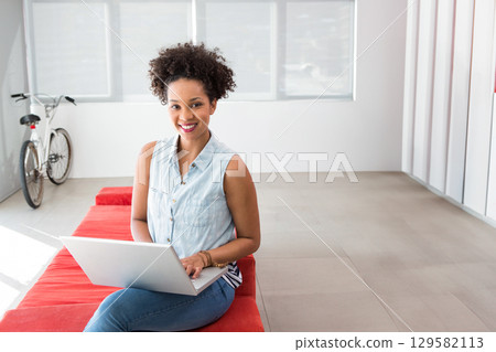 African American female sitting on red bench using silver laptop in lounge with bicycle and blinds 129582113