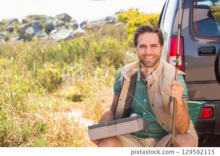 Man sitting on chair at SUV tailgate holding fishing rod, tackle box in meadow, copy space 129582115
