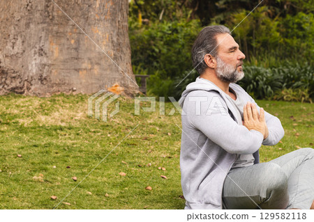 Mature adult man meditating on grass in park wearing hoodie and sweatpants by tree, copy space Mature adult man meditating on grass in park wearing hoodie and sweatpants by tree, copy space 129582118