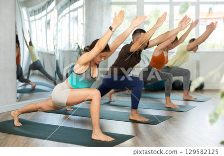 Lovers of active lifestyle on yoga mat perform doing version of warrior pose in gym Lovers of active lifestyle on yoga mat perform doing version of warrior pose in gym 129582125