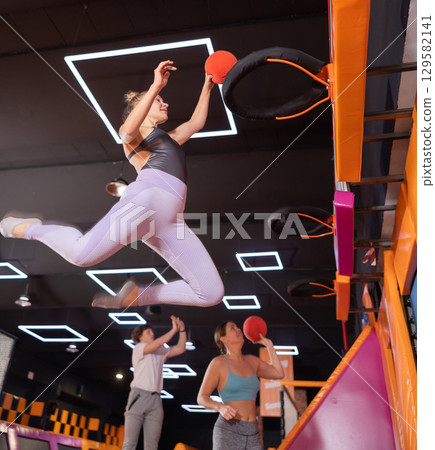 Middle-aged woman throwing ball in basket high jumping in trampoline arena Middle-aged woman throwing ball in basket high jumping in trampoline arena 129582141