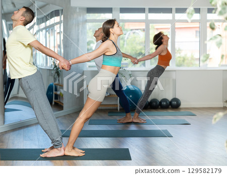 Friendly sporty couple practice yoga poses, standing back to back and holding each other from back, keep balance during group acro yoga class in yoga studio Friendly sporty couple practice yoga poses, standing back to back and holding each other from back, keep balance during group acro yoga class in yoga studio 129582179