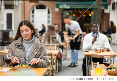 Young woman sitting on the terrace of a restaurant and eating salad 129582420