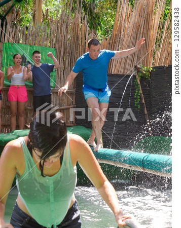 Concentrated man crossing pool of water on inflatable log with hanging obstacles Concentrated man crossing pool of water on inflatable log with hanging obstacles 129582438