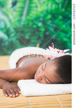African American woman reclining on bamboo mat spa table outside wearing pink flower and towel 129582525