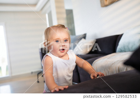 Female infant standing and leaning against dark gray sofa cushions in living room by window blinds Female infant standing and leaning against dark gray sofa cushions in living room by window blinds 129582533