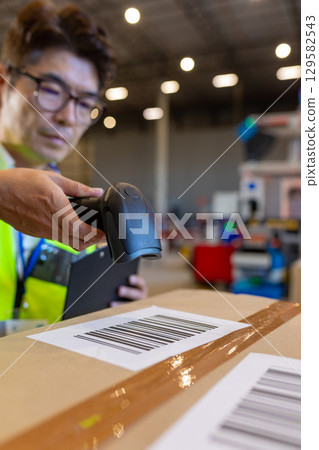 Asian man in safety vest scanning barcode on box and holding clipboard in warehouse, copy space Asian man in safety vest scanning barcode on box and holding clipboard in warehouse, copy space 129582543