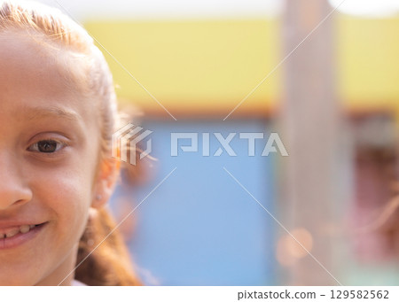 Girl smiling and gazing at camera by wooden post near pastel walls in courtyard, copy space 129582562