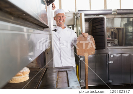 Mature male baker sliding loaf from industrial oven with wooden peel in commercial bakery kitchen Mature male baker sliding loaf from industrial oven with wooden peel in commercial bakery kitchen 129582587