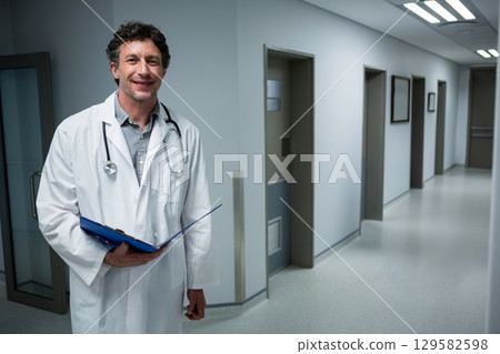 Senior male doctor wearing coat and stethoscope holding clipboard in hospital hallway, copy space Senior male doctor wearing coat and stethoscope holding clipboard in hospital hallway, copy space 129582598